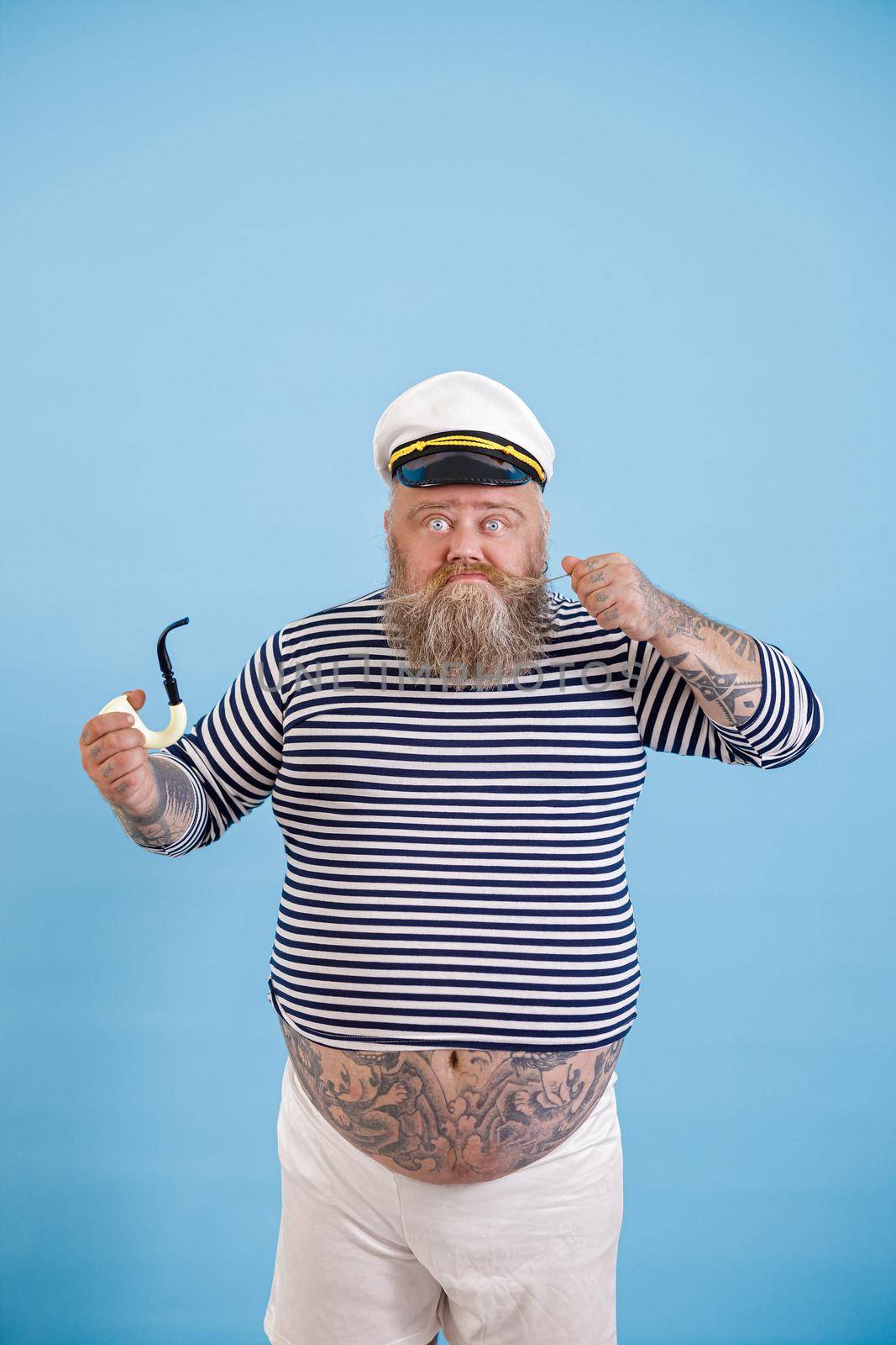 Funny bearded obese man wearing sailor suit with smoking pipe tightens up mustache on light blue background in studio