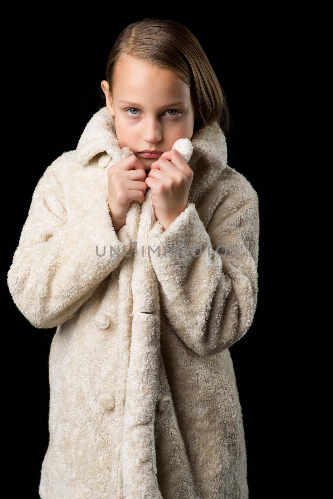 Portrait of girl in beige faux fur coat. Beautiful fashionable blonde girl keeping hands on her collar posing against black background. Trendy child dressed fashion, stylish outerwear
