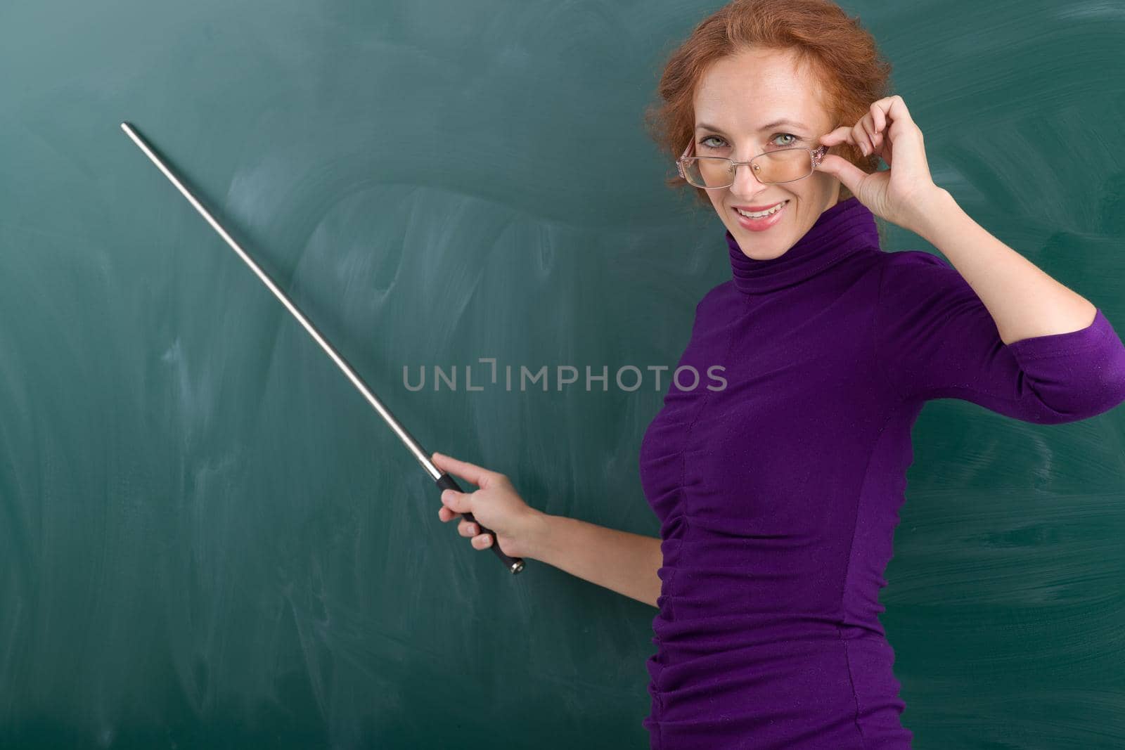 Teacher standing at blackboard with pointer. Portrait of smiling young woman looking over glasses. Teacher posing at green chalkboard and pointing at it