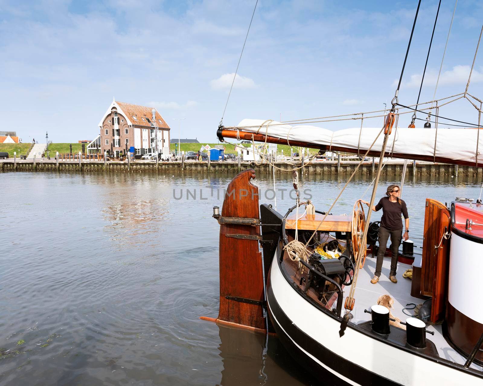 oudeschild, netherlands, 19 july 2021: woman steers old wooden sailing vessel in port of oudeschild on dutch island of texel while boys fish for crabunder blue sky in the netherlands