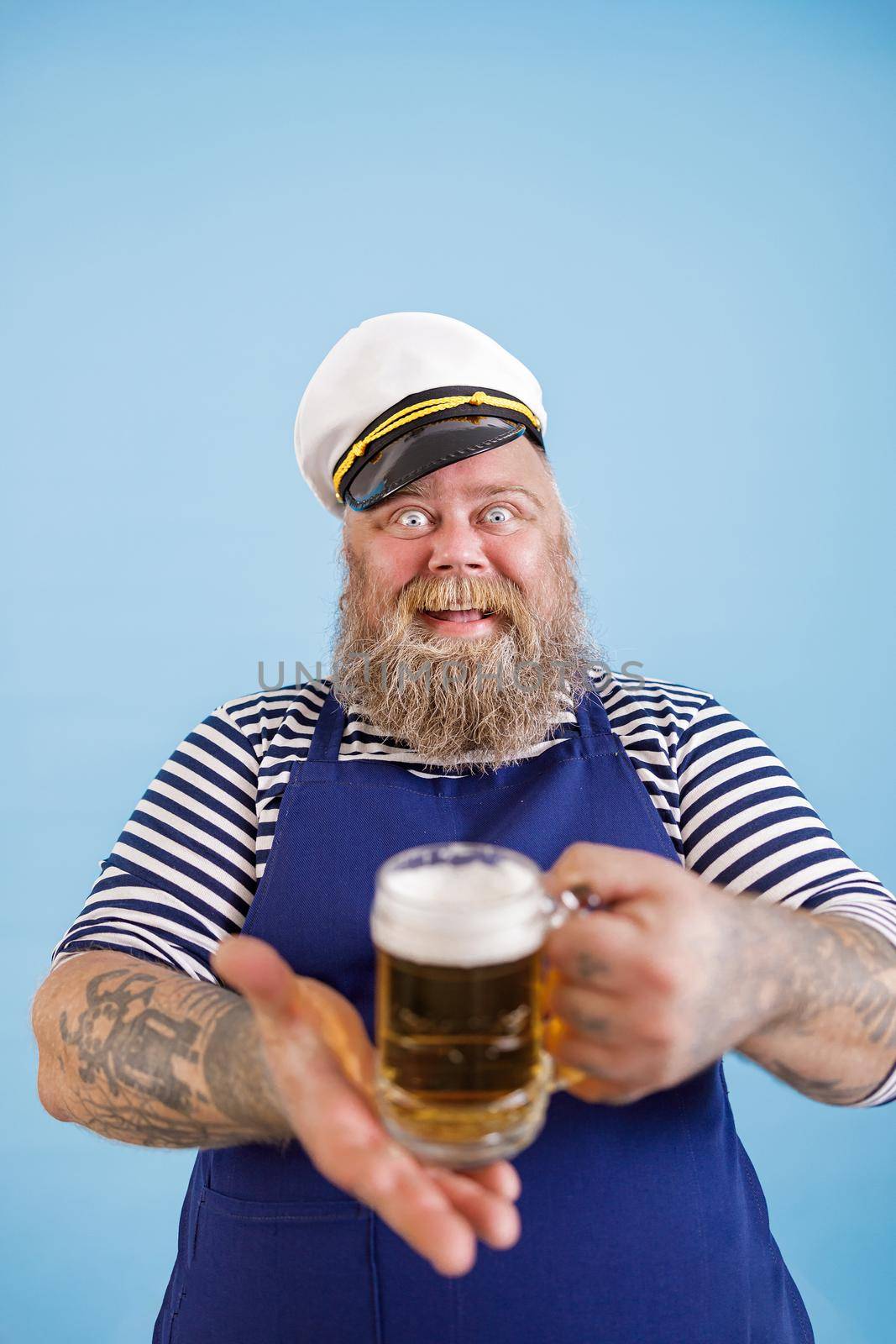Cheerful mature plump man in sailor costume with apron and captain hat offers glass mug of delicious beer on light blue background in studio