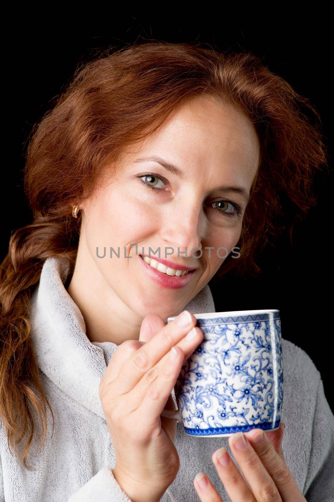 Woman holding mug in her hands. Cheerful brown haired woman in warm sweatshirt posing with cup of coffee or tea against black background. Female person enjoying of hot drink in the morning