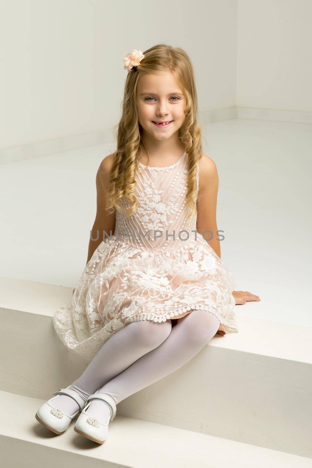 Lovely girl sitting on white staircase. Portrait of beautiful blonde girl wearing nice dress, tights and shoes posing in studio. Lovely smiling seven years old kid sitting on white stairs
