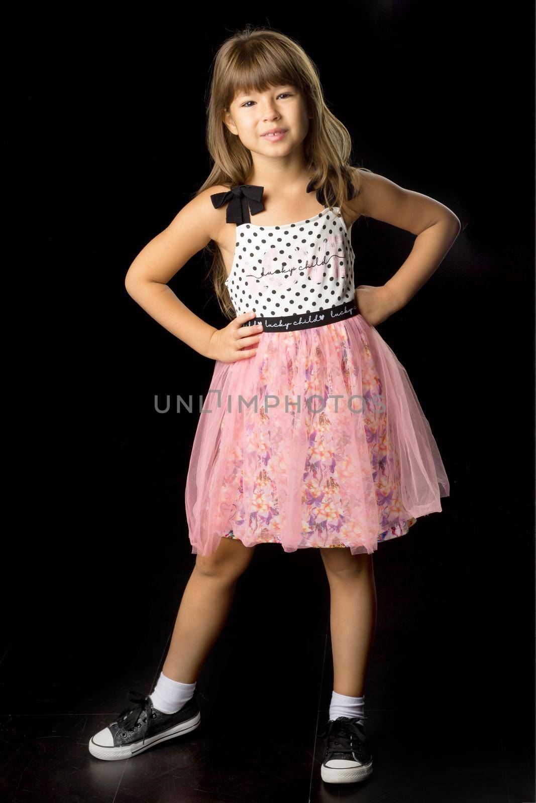 Portrait of cheerful girl in stylish summer clothes. Front view of beautiful girl wearing sundress and headband posing with hand on her waist against black background and smiling at camera