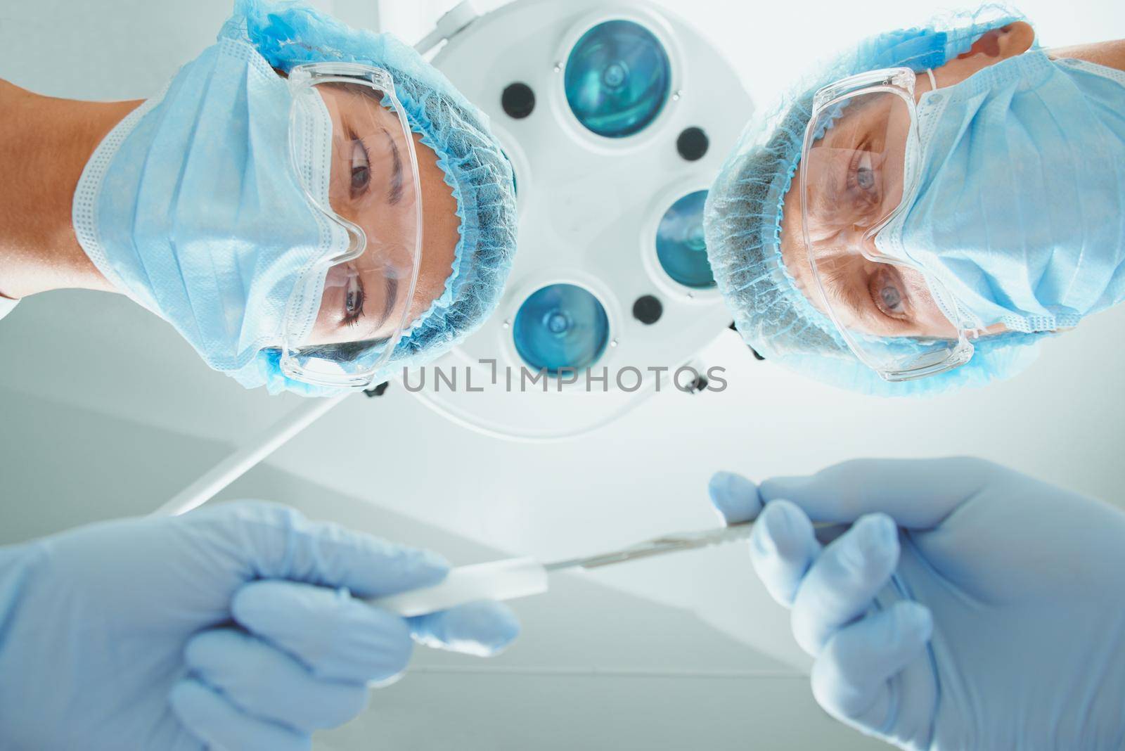 Female nurse passes a scalpel to man surgeon on operation on background of surgical lamp