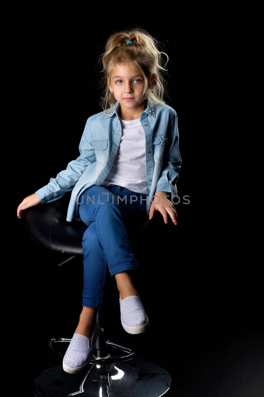 Smiling blonde girl sitting on a high chair in the studio, sitting with her head bowed down, portrait of a cute girl isolated on black background