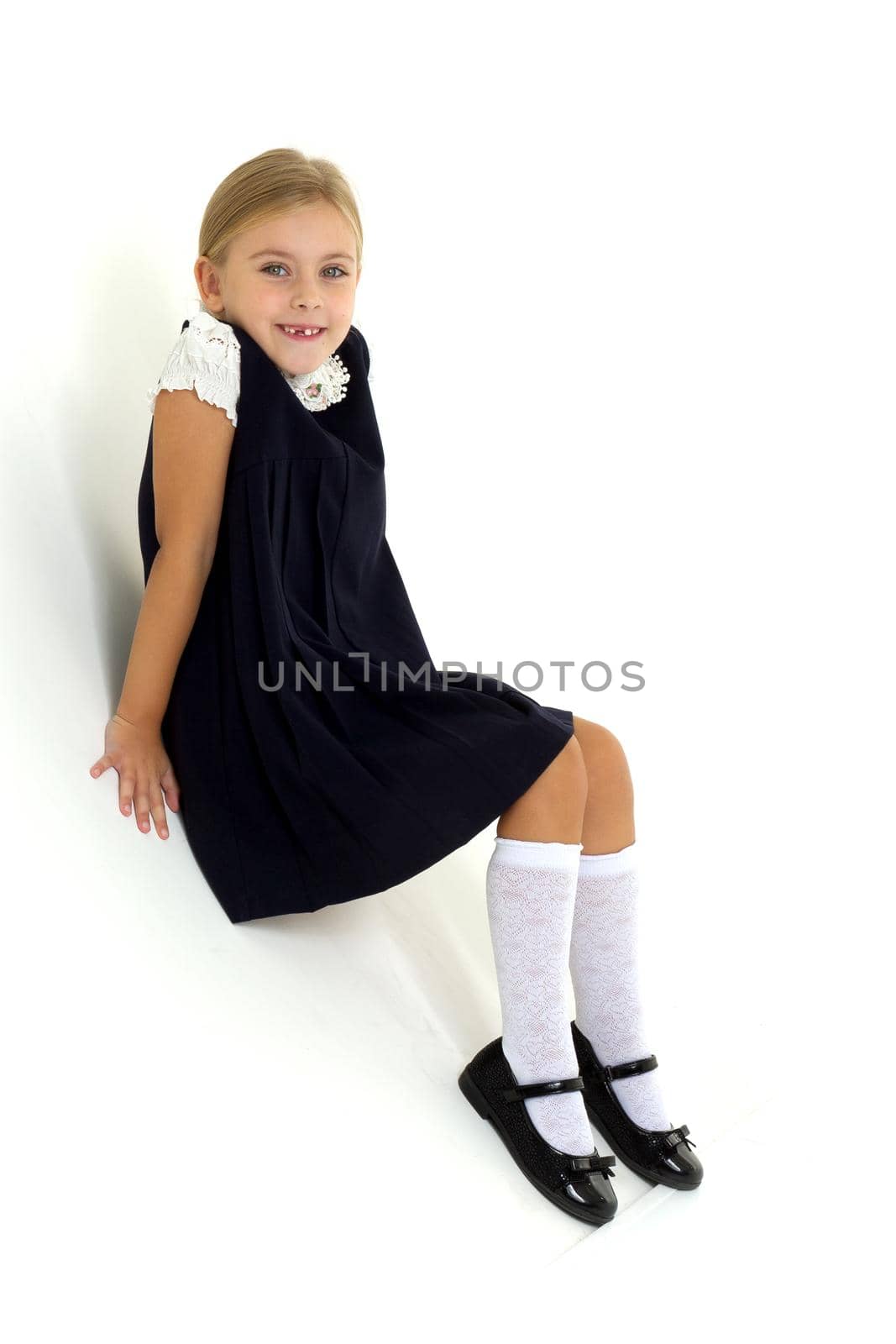 Lovely girl posing in studio on background. Cheerful blonde girl wearing school uniform sitting on floor leaning back on her hands. Cute seven years old kid in white blouse and blue dress