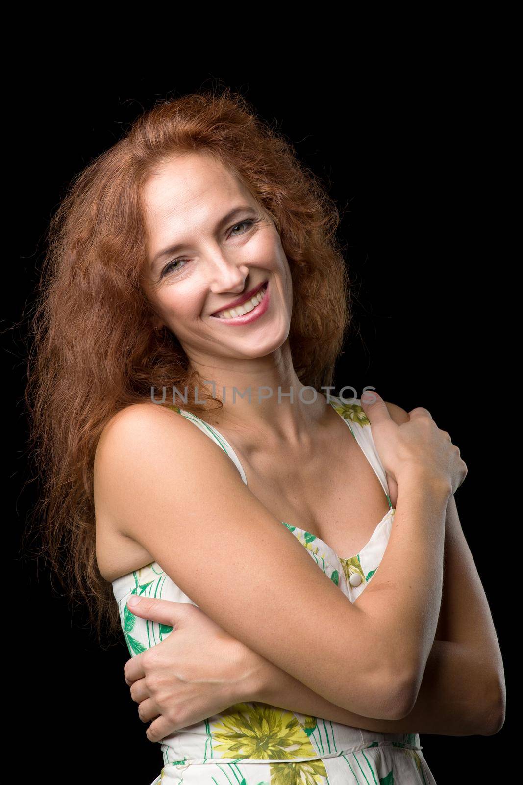 Beautiful smiling woman hugging herself. Cheerful young woman in floral sundress standing against black background. Portrait of dreaming girl posing in studio