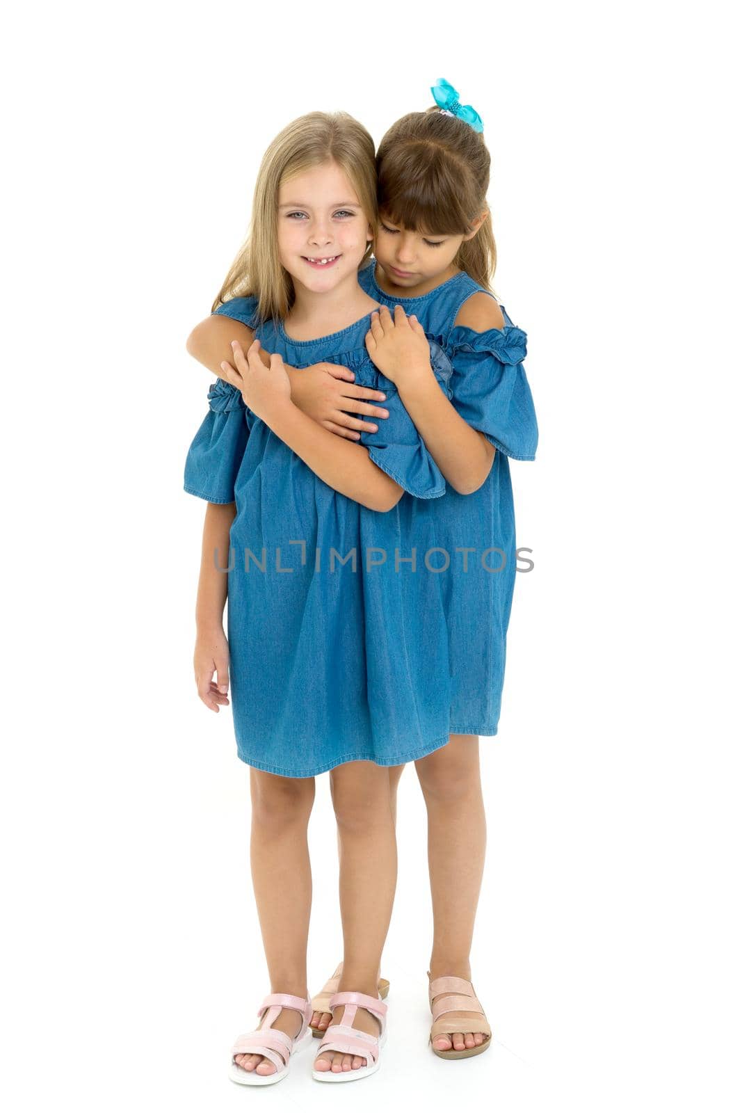 Portrait of a cute girl. Adorable girls dressed in matching denim dresses hug each other tightly with their backs pressed against an isolated white background.