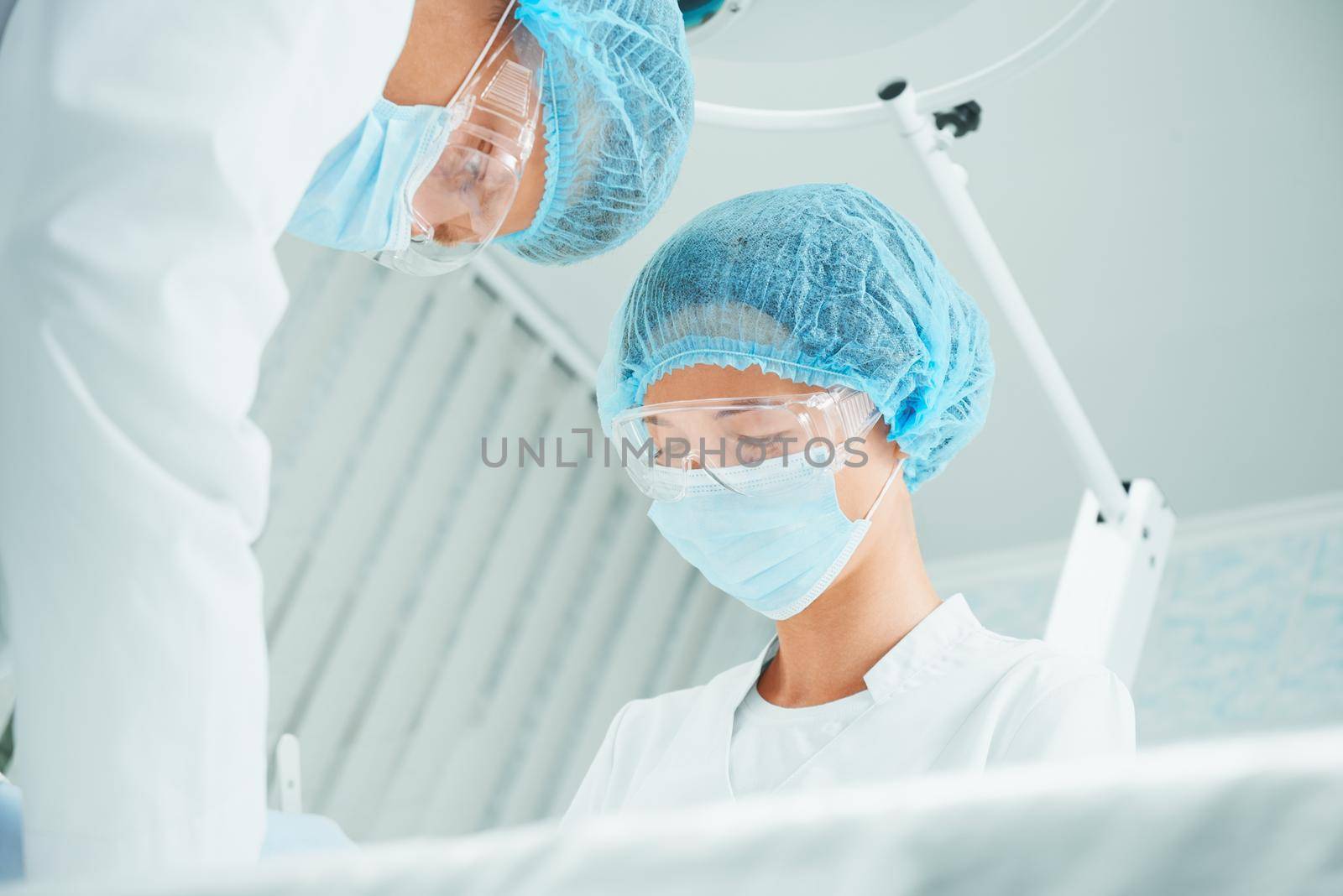 Male and female serious surgeons are working in operating room on background of surgical lamp, teamwork