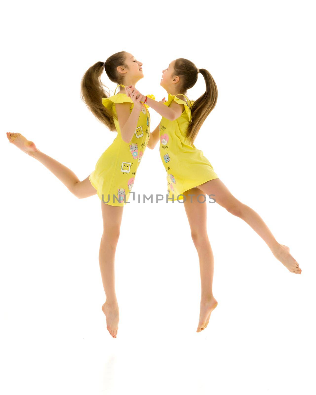 Side View of Barefoot Teen Girls Looking at Each Other Standing on One Leg on Toes, Two Beautiful Sisters Wearing Yellow Short Dresses, Full Length Portrait of Teenagers Isolated on White Background.