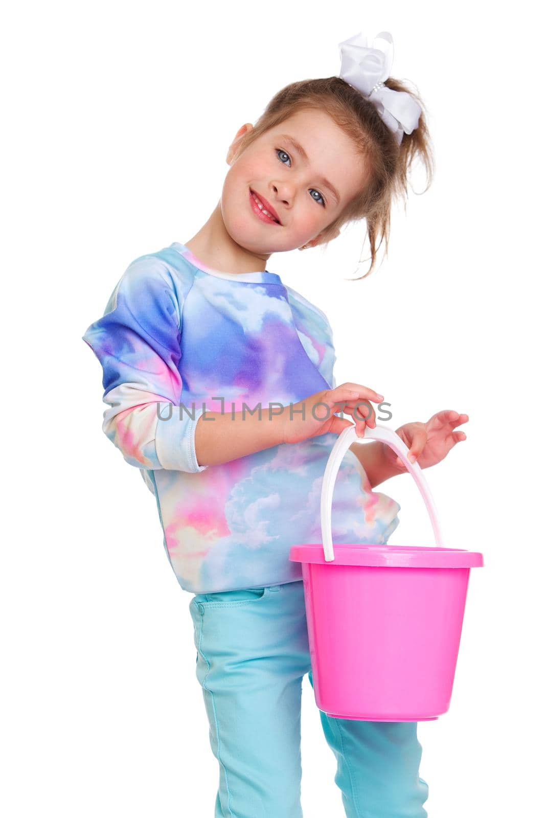 Lovely little girl standing with bucket. Cute happy kid dressed casual clothes holding pink bucket against white background. Portrait of adorable playful girl posing in studio