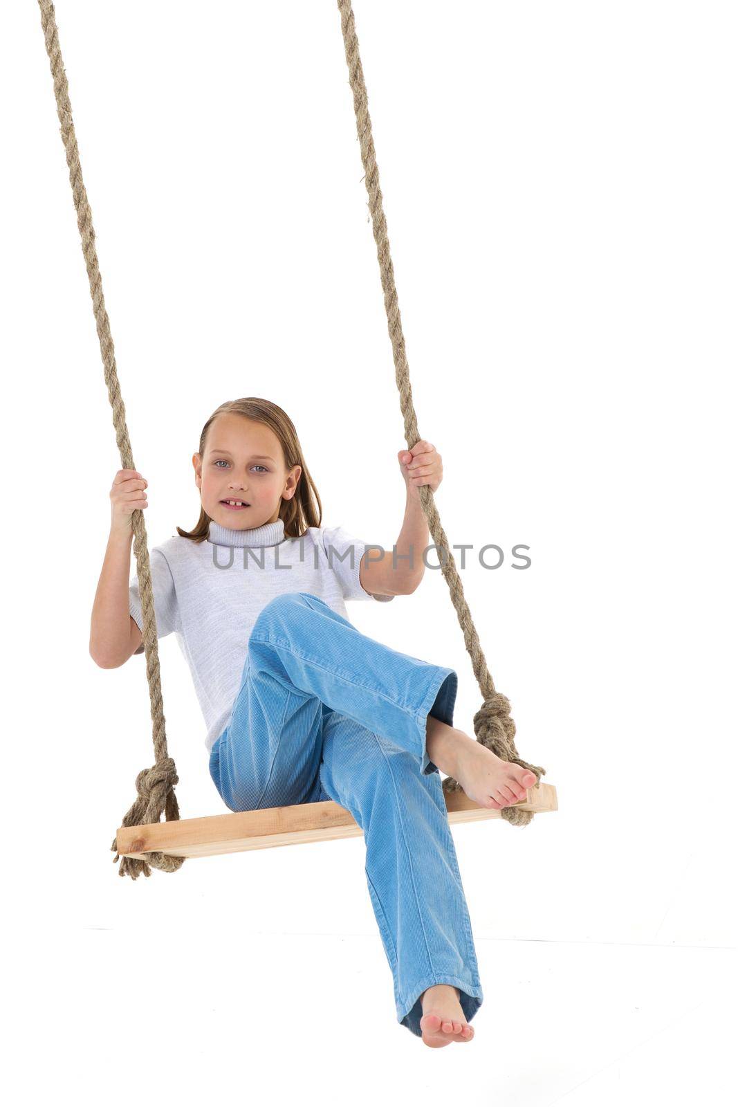 Blonde girl swinging on rope swing. Full length shot of pretty stylish barefoot girl having fun against white background. Portrait of preteen child posing in studio