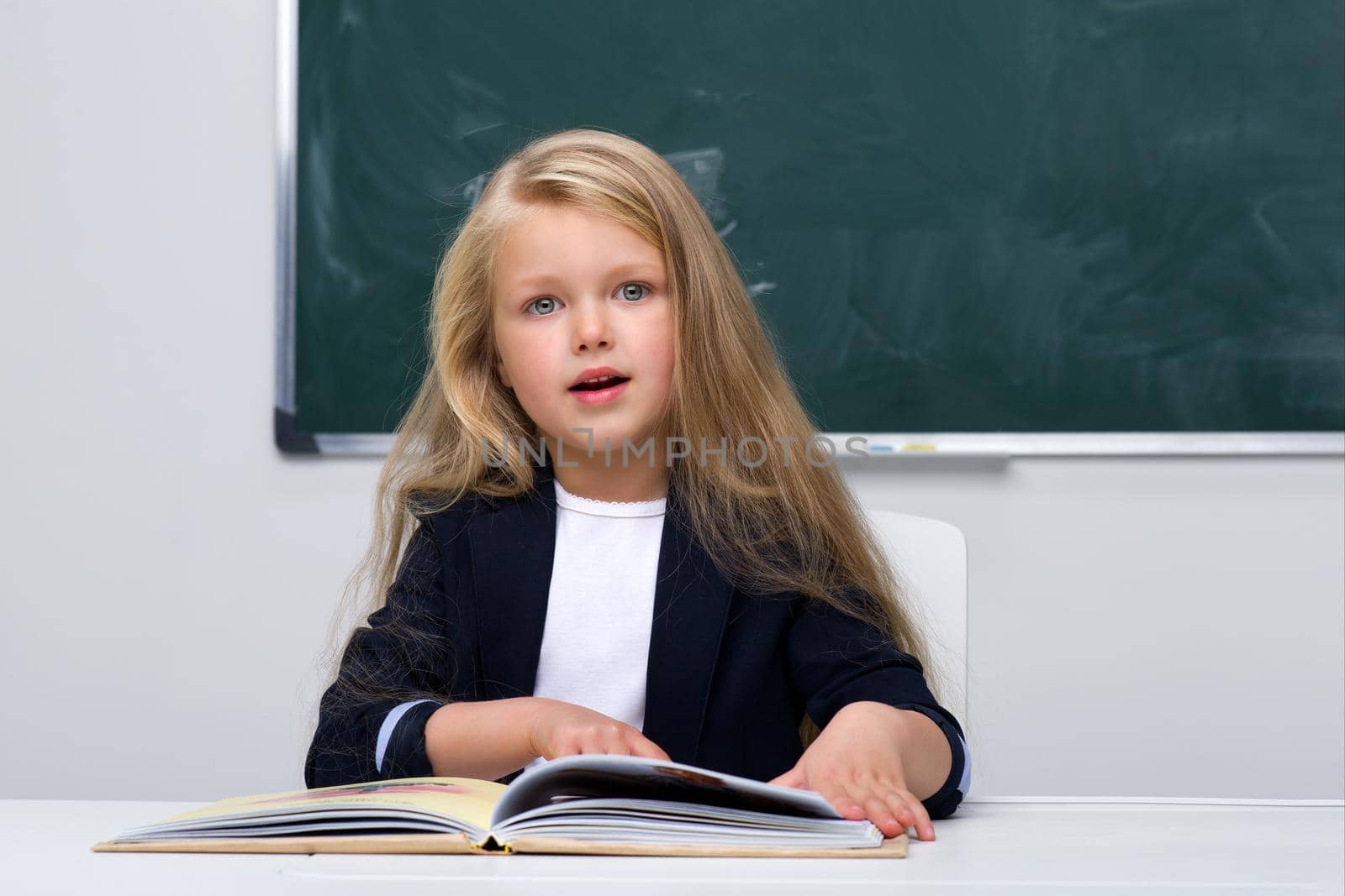 Happy schoolgirl sitting at desk. Elementary school student in blue jacket and white blouse reading book on background of blackboard in classroom. Back to school, education concept