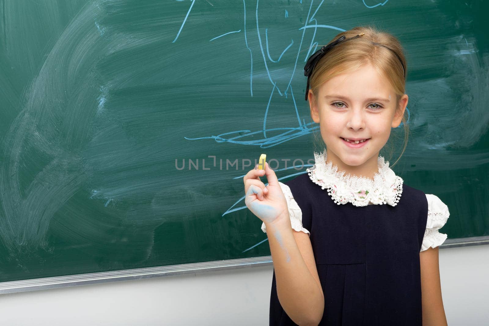 Cute blonde girl standing at green chalkboard. Portrait of beautiful smiling schoolgirl in uniform standing in front of blackboard. School and education concept