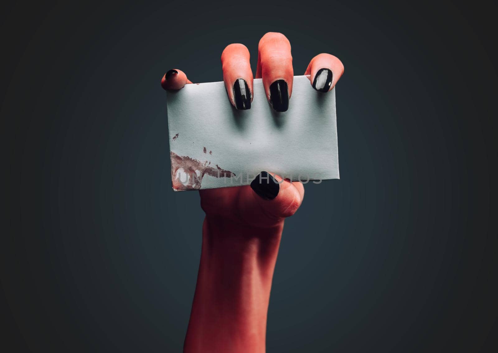 Red demon hand holds empty white card on dark background, space for text. Halloween or horror theme