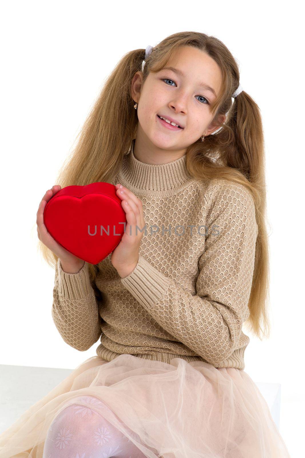 Beautiful girl holding red heart in her hands. Cute preteen girl with pigtails wearing knitted jumper sittingon cube and looking at camera against white background.