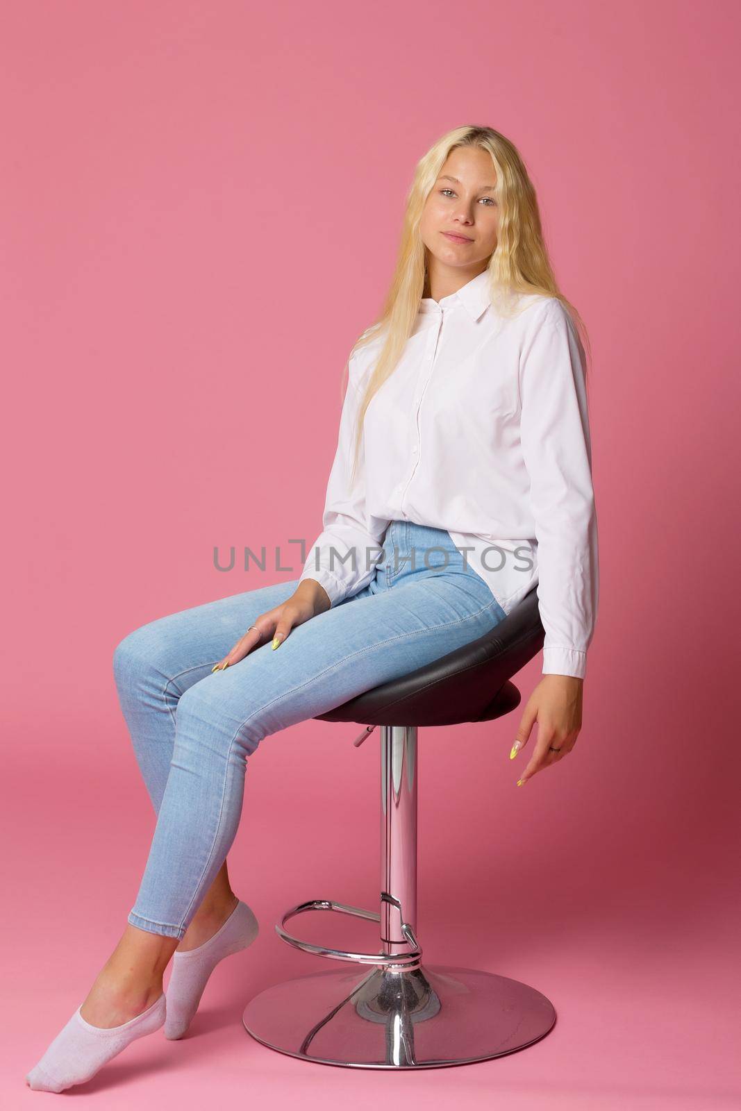 Smiling blonde girl sitting on a high chair in the studio, sitting with her head bowed down, portrait of a cute girl isolated on Pink background