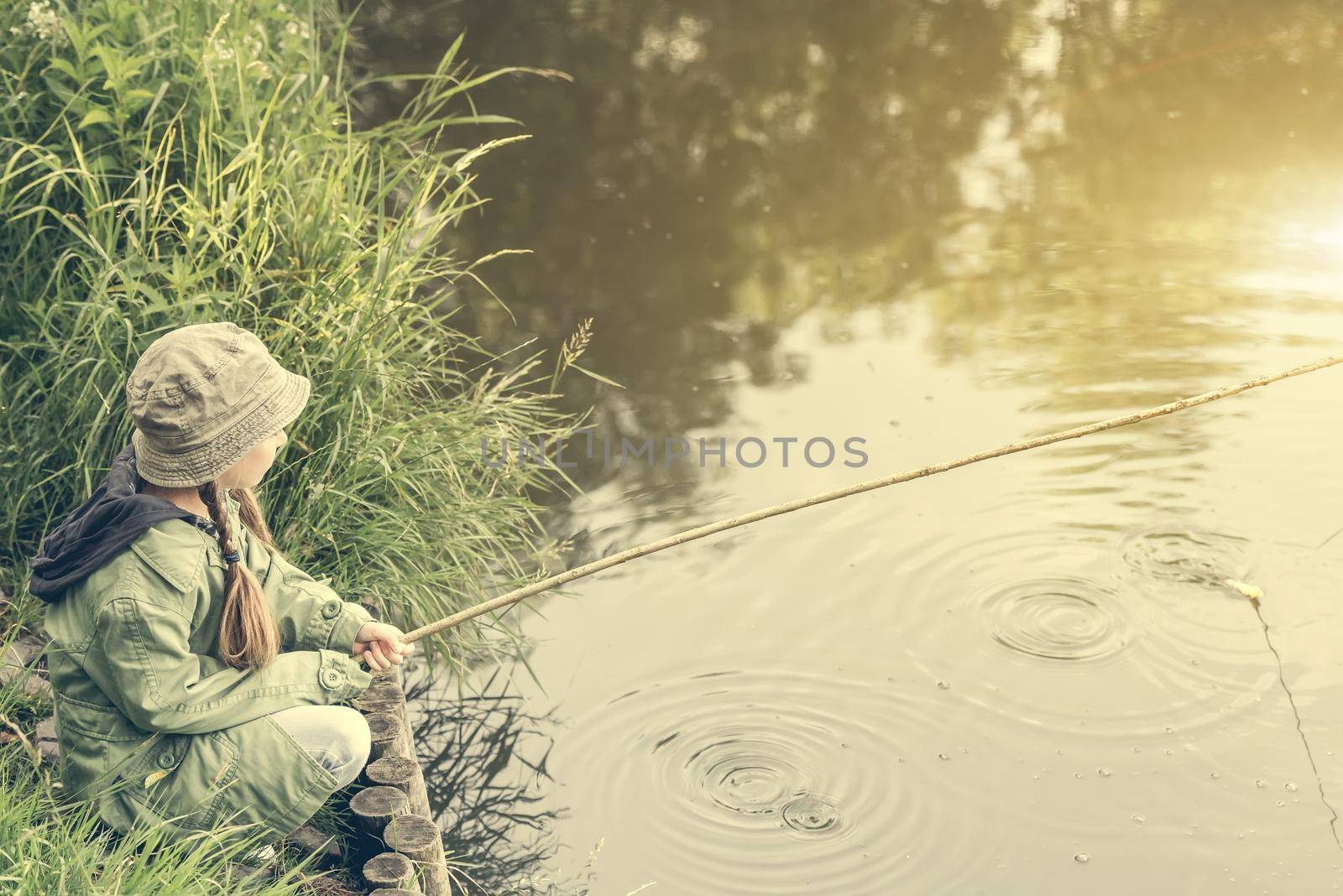 little fisher girl sitting on a river bank with a rod