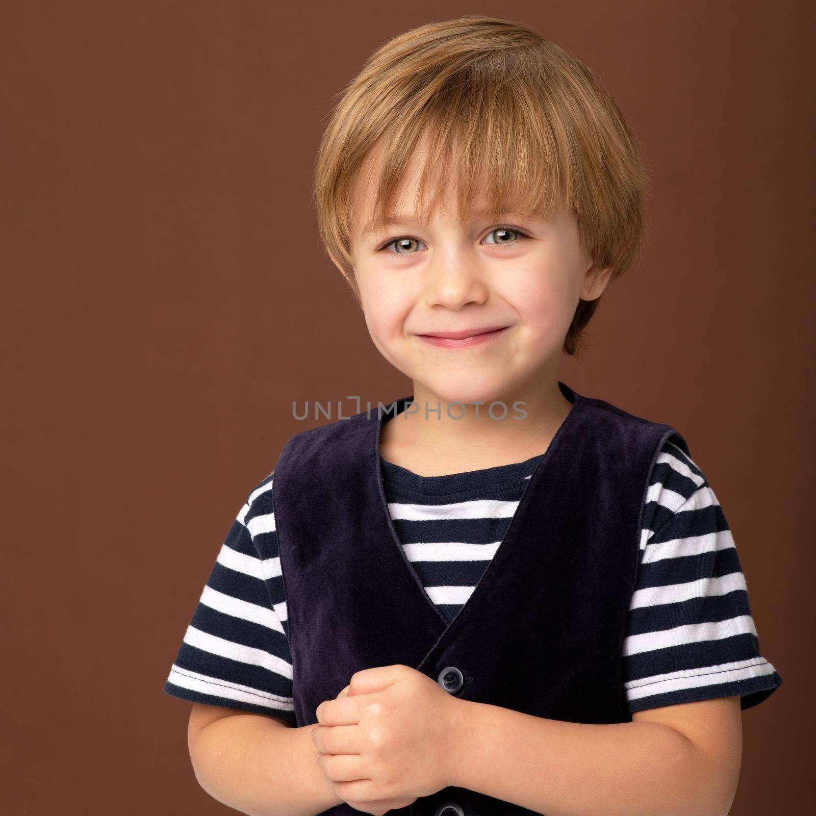 Portrait of happy joyful little boy. Lovely smiling kid wearing striiped t-shirt and blue velvet vest standing on brown background. Cheerful little boy in stylish trendy clothes