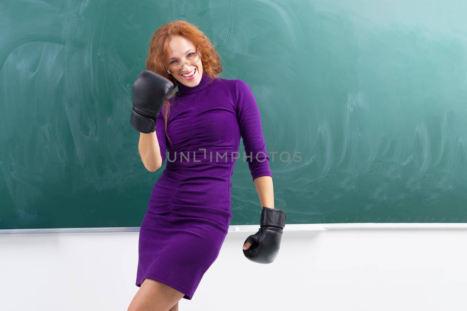 Happy teacher wearing black boxing gloves standing in front of blank blackboard. Young woman wearing glasses and purple casual dress enjoying her victory