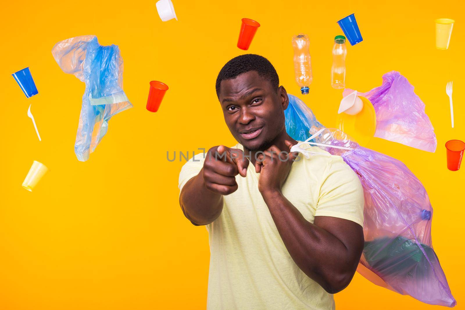 Problem of trash, plastic recycling, pollution and environmental concept - funny african american man carrying garbage for recycling and pointing on you on yellow.