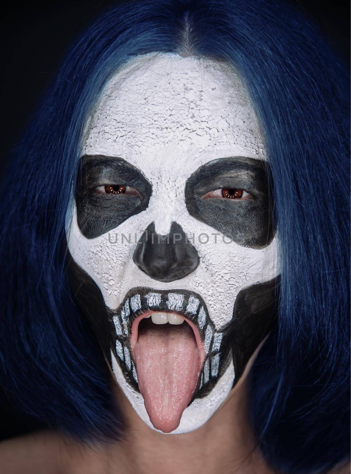 Woman with skull makeup and blue hair showing tongue on dark background, Halloween or horror theme