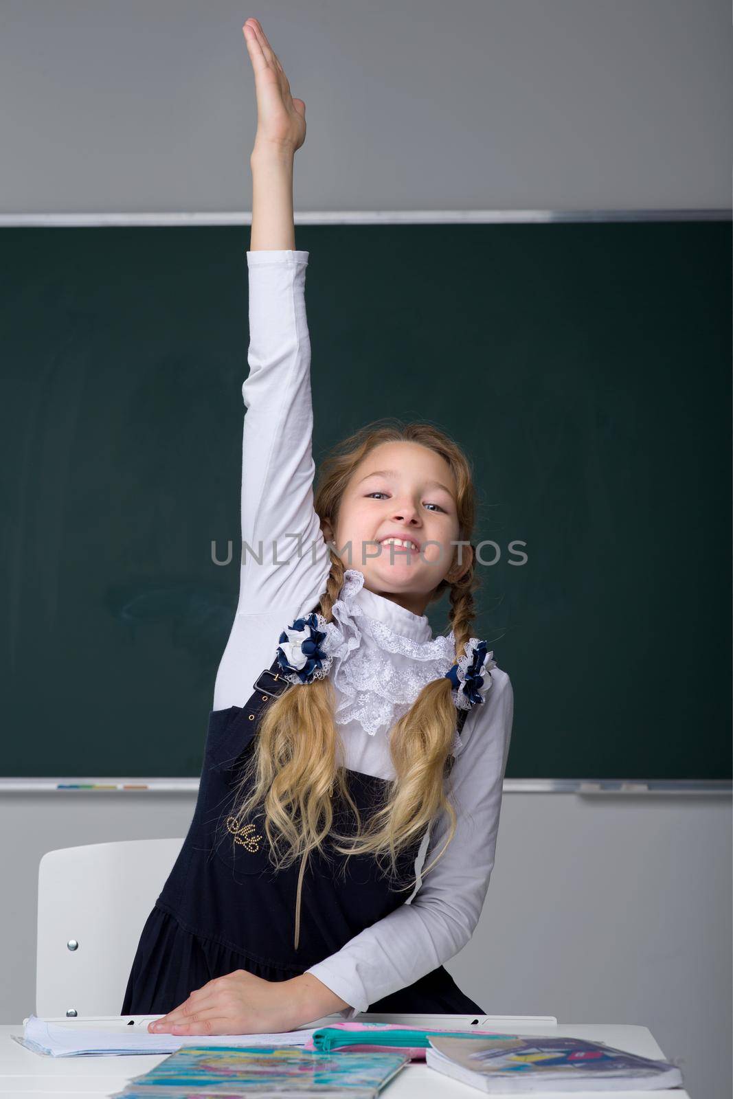 Schoolgirl learning in classroom. Smiling girl in uniform rising her hand up to answer the teacher. Primary school student sitting on background of blackboard. Back to school, education concept