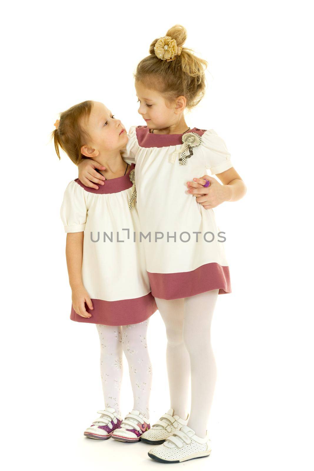 Cute adorable little sisters standing and hugging. Lovely girls dressed in the same dresses looking at each other against white background. Two happy kids embracing each other