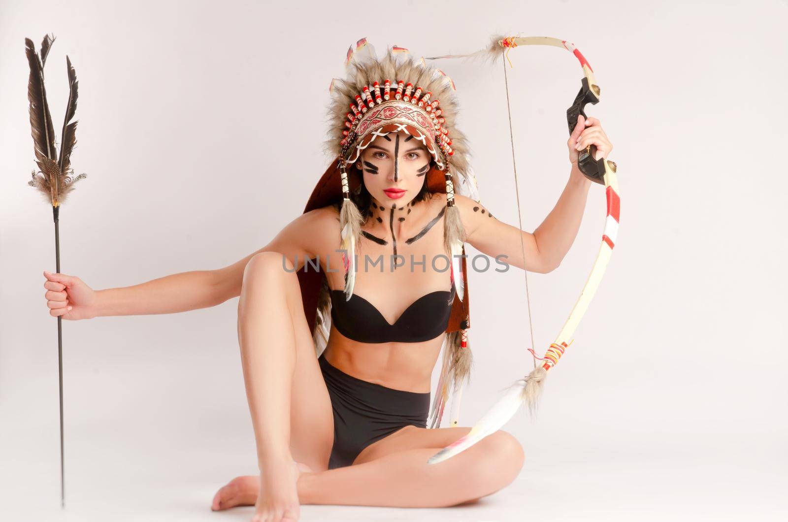 a woman in the image of indigenous peoples of America with a bow and arrow poses sitting on a light background by Rotozey