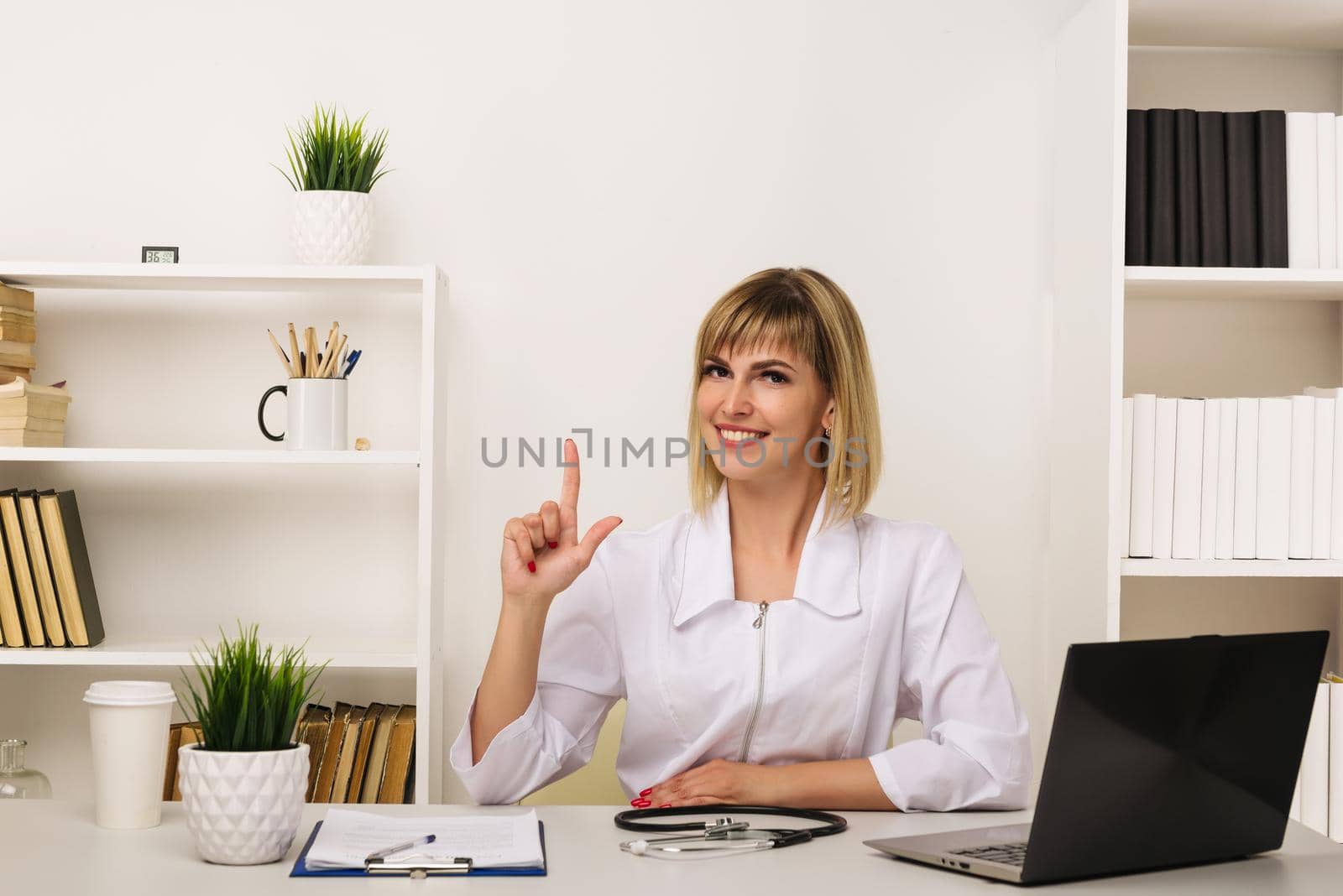 Friendly female doctor works at her desk in the office pointing up - image