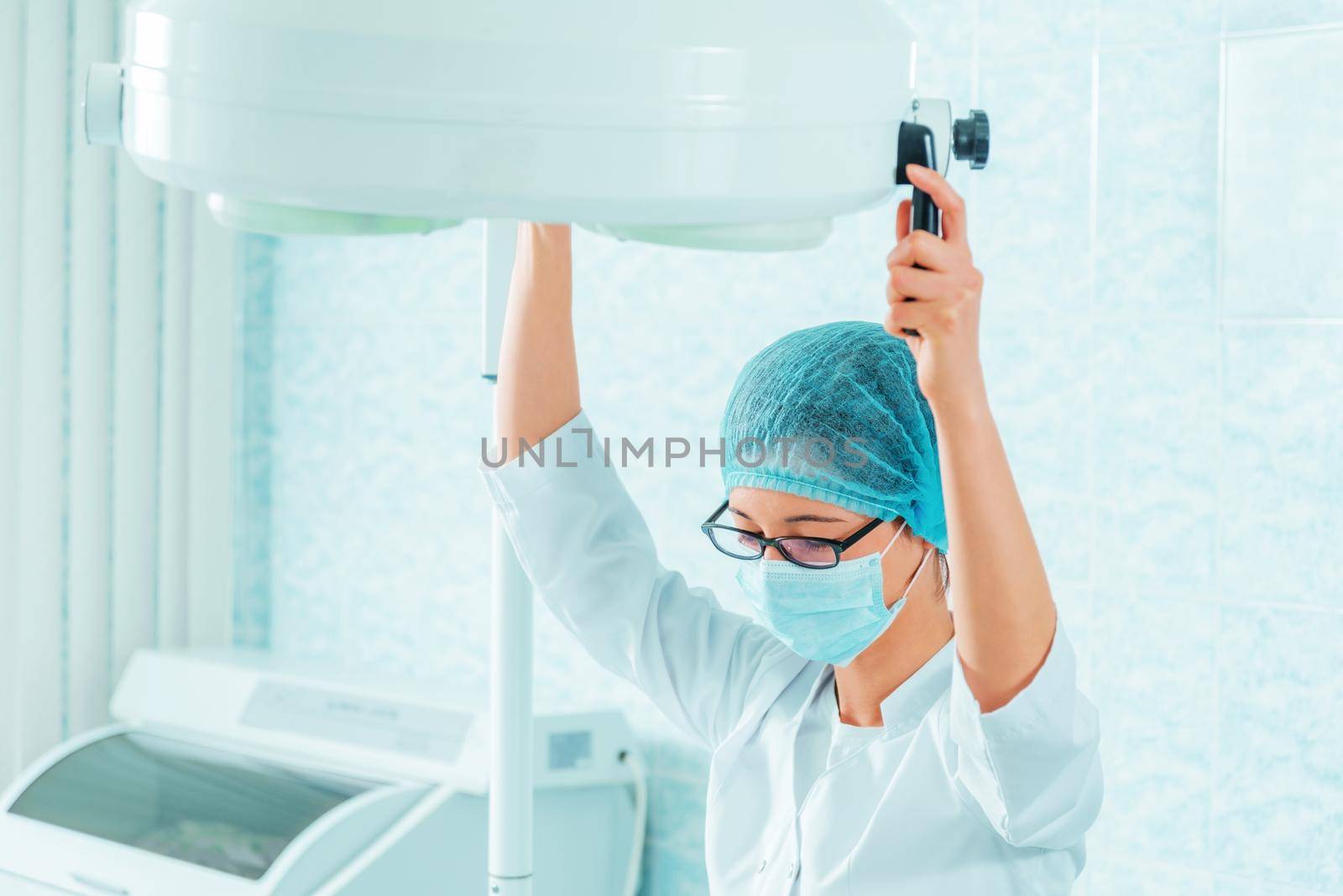 Medical assistant in a mask and cap places a surgical lamp for operation
