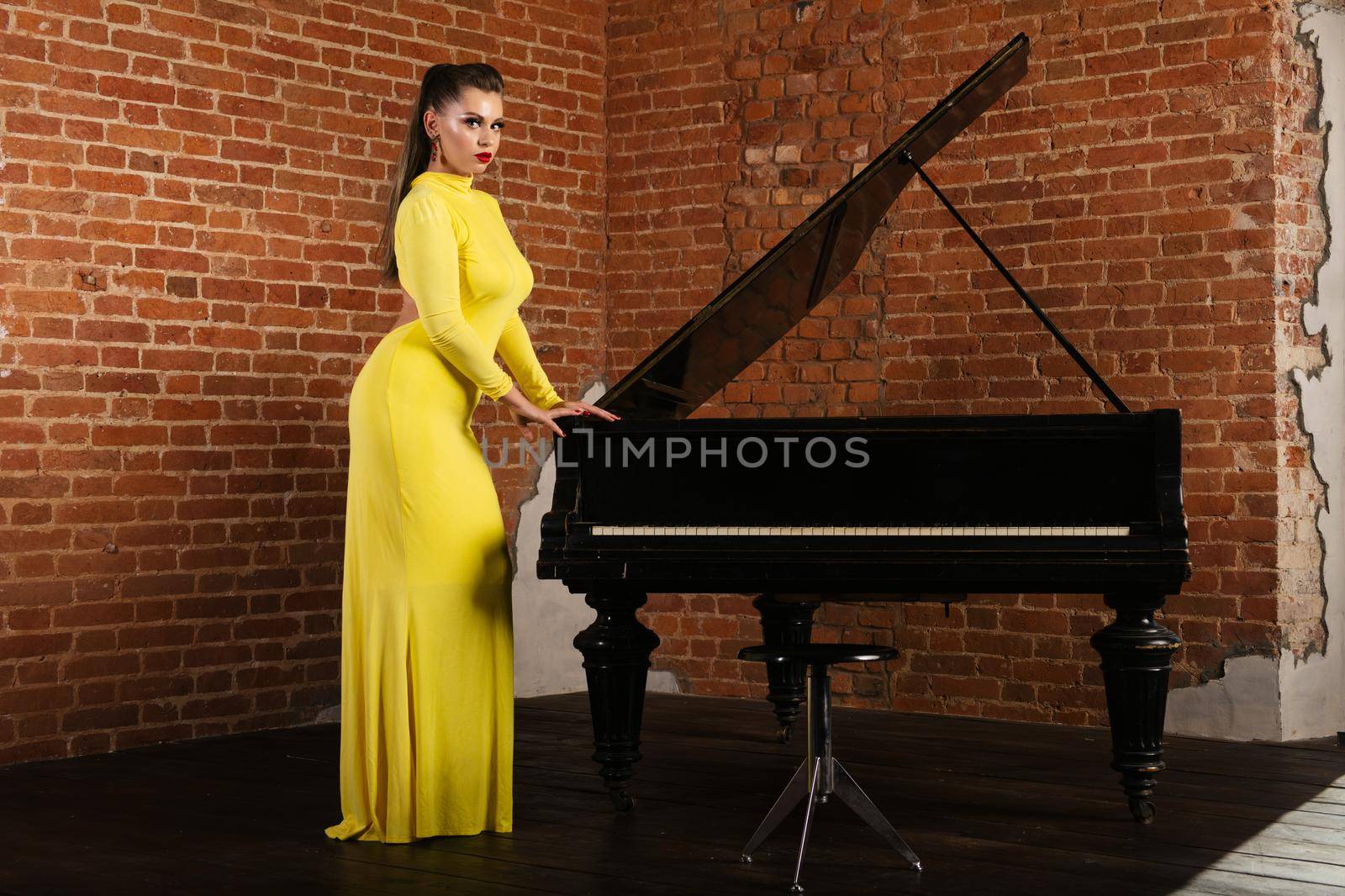 Beautiful sensual and sexy blonde girl in a yellow dress standing near the piano against a brick wall