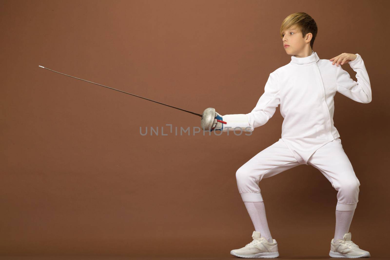 Boy fencer standing in attacking pose. Portrait of teenage boy wearing white fencing suit posing with sabre against brown background. Teenager taking part at foil fencing tournament