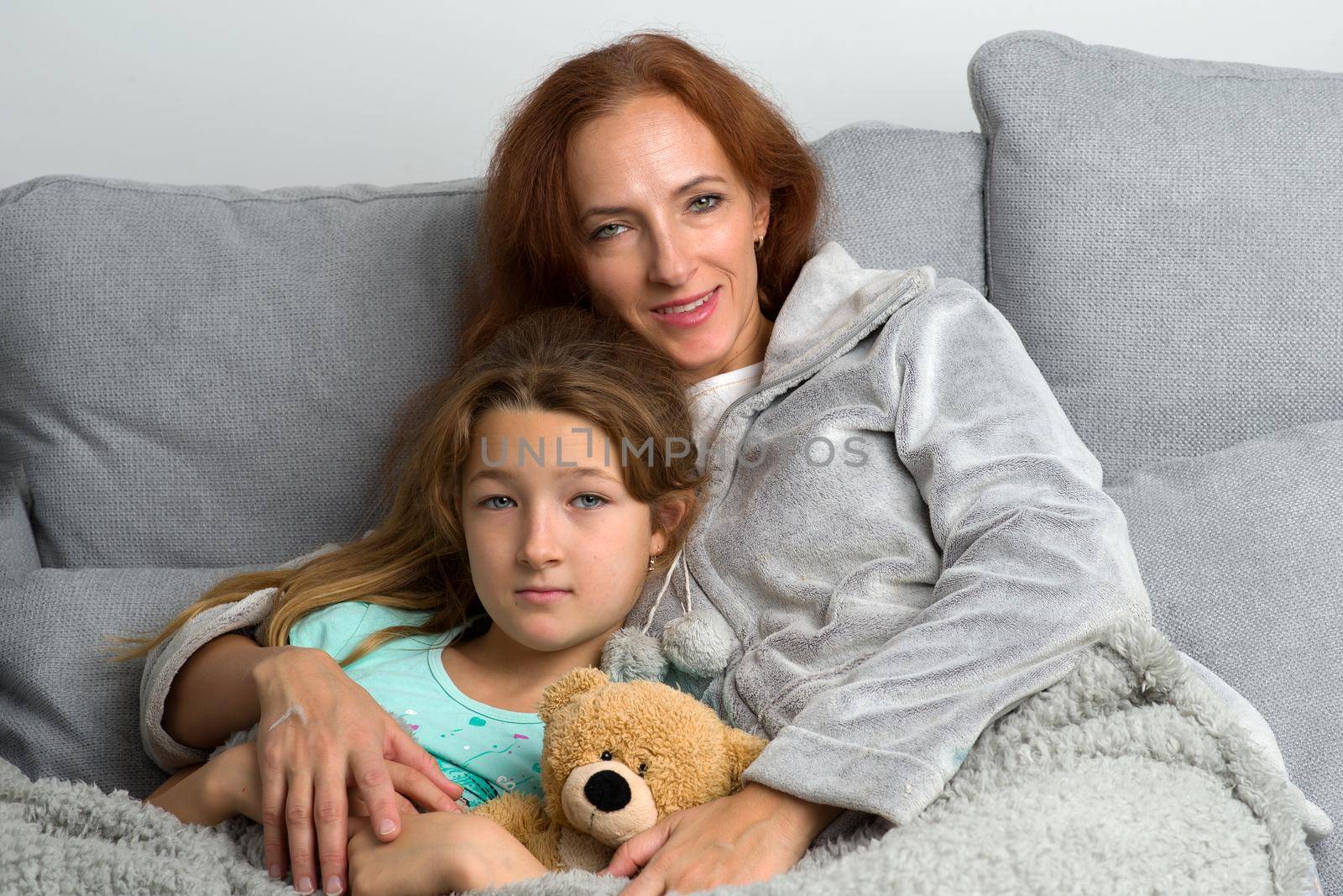 Happy mom sitting on couch with her preteen daughter. Smiling loving mother hugging her daughter lying with teddy bear toy. Woman and child wearing indoor clothes sitting together covered with plaid