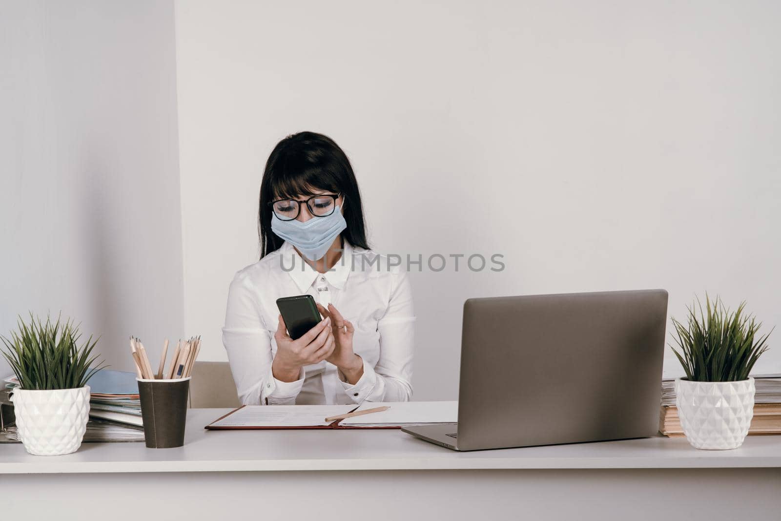 A young woman works remotely in the office with a protective mask during an epidemic. Looks at the message in the smartphone