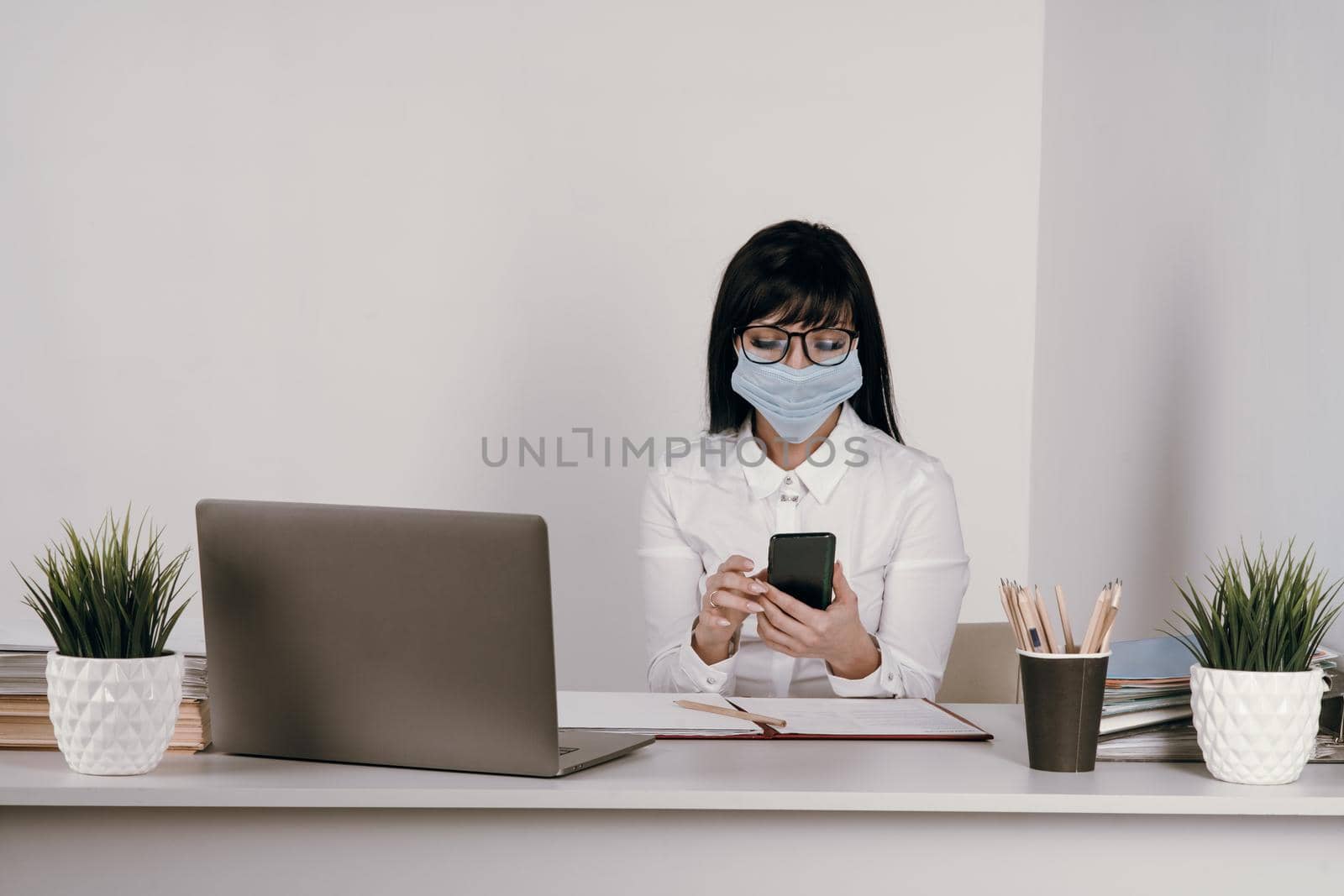 A young woman works remotely in the office with a protective mask during an epidemic. Looks at the message in the smartphone