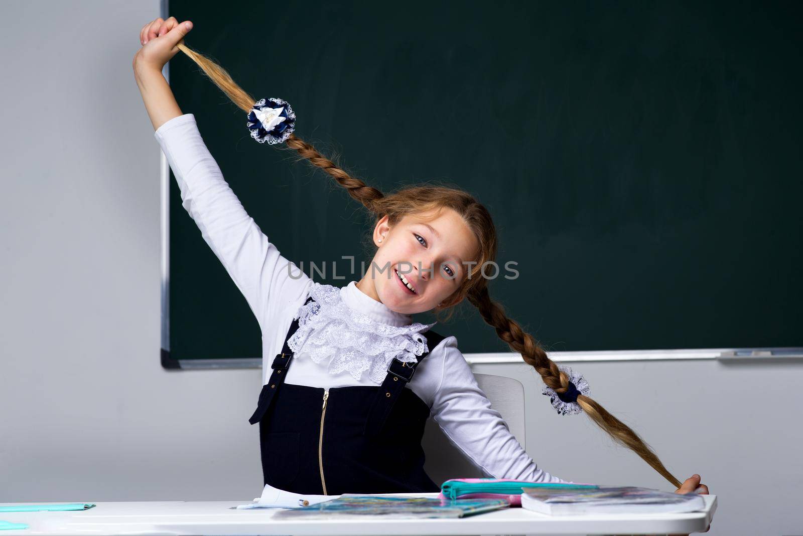 Schoolgirl holding her braids. Cheerful primary school student in uniform sitting on background of blackboard in classroom. Back to school, education concept