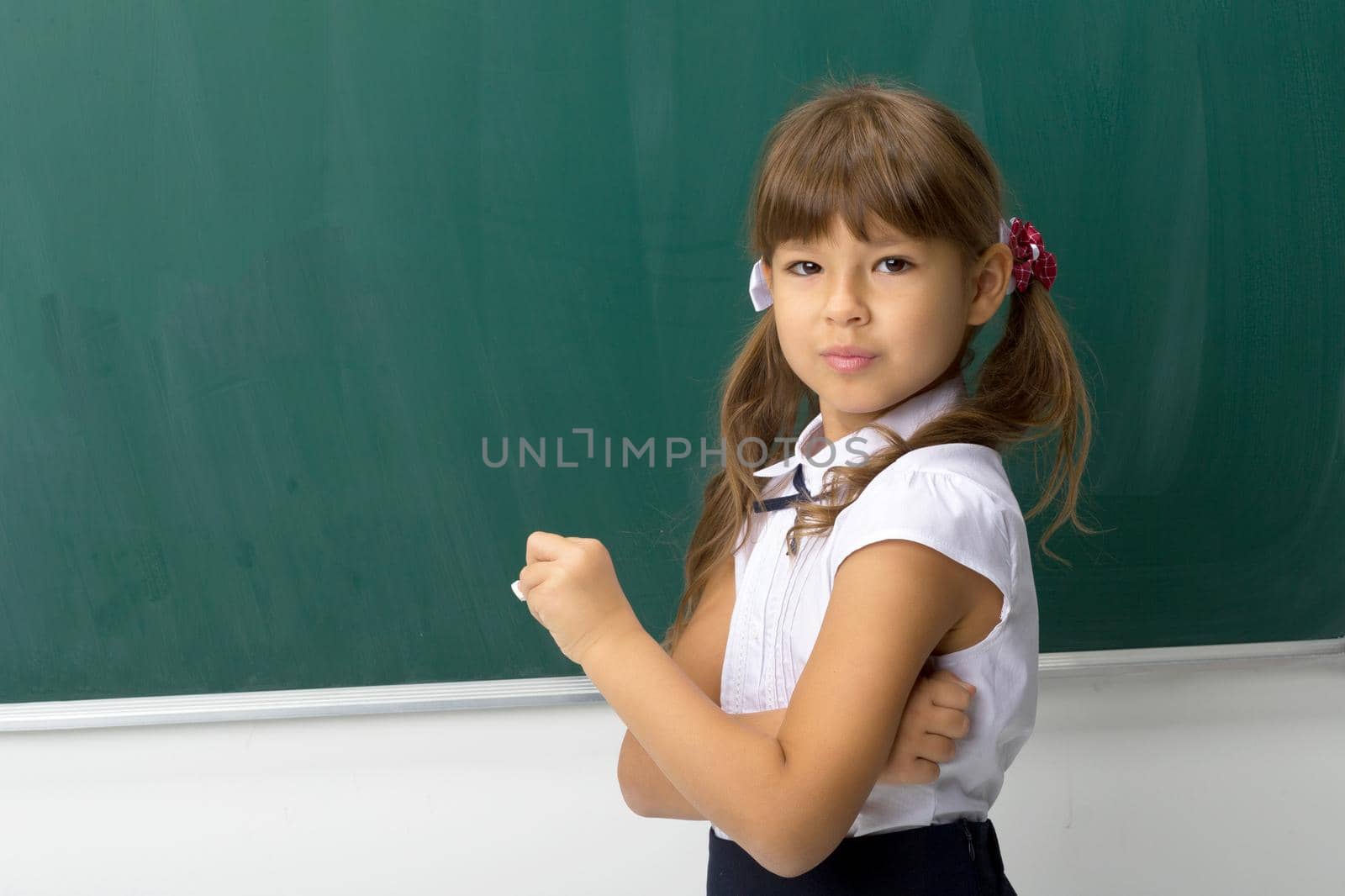 Pretty girl posing at blackboard. Portrait of beautiful schoolgirl in school uniform standing at green chalkboard with piece of chalk in her hand and looking at camera. School and education concept
