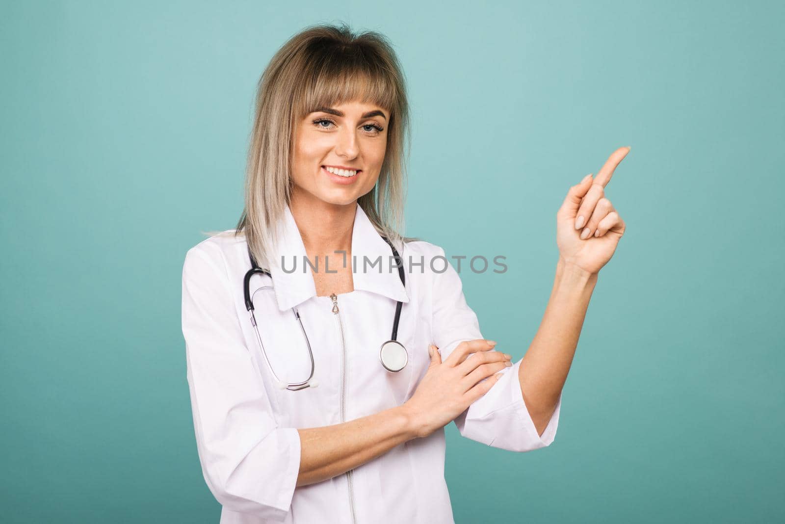 Smiling young female doctor with a stethoscope points up her fingers on a blue background