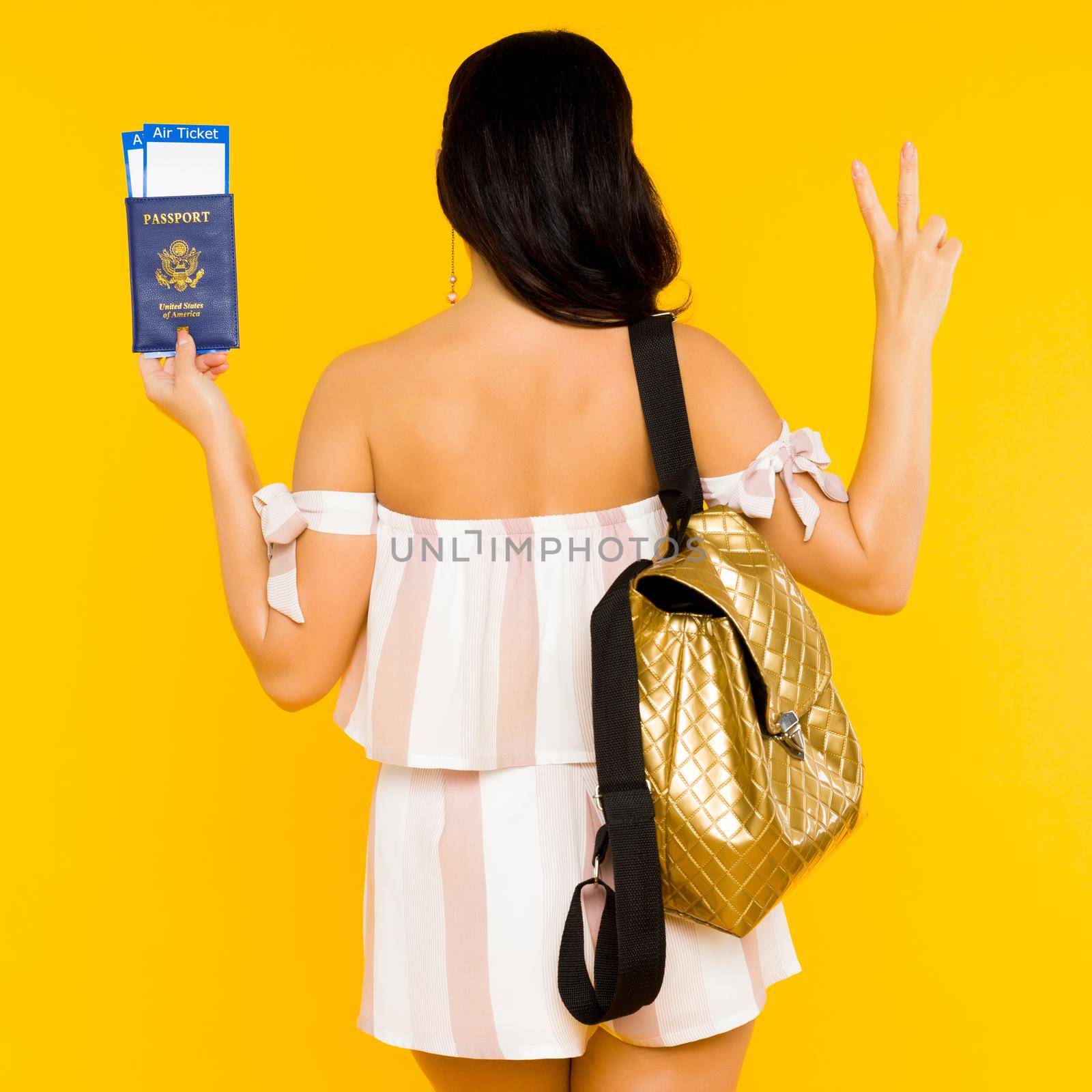 Travel concept. Young asian woman holding passport with tickets standing back with knapsack shows peace sign on yellow background