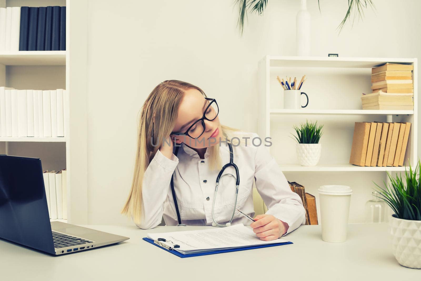 Young tired exhausted woman sitting at desk, working on computer with medical documents in light office in hospital. Female doctor in medical gown sleep in consulting room. toned