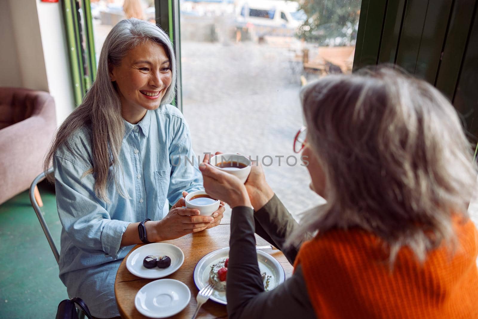 Pretty happy mature women companions with natural grey hair hold cups of hot drinks sitting at small table in cozy cafe. Long-time friendship