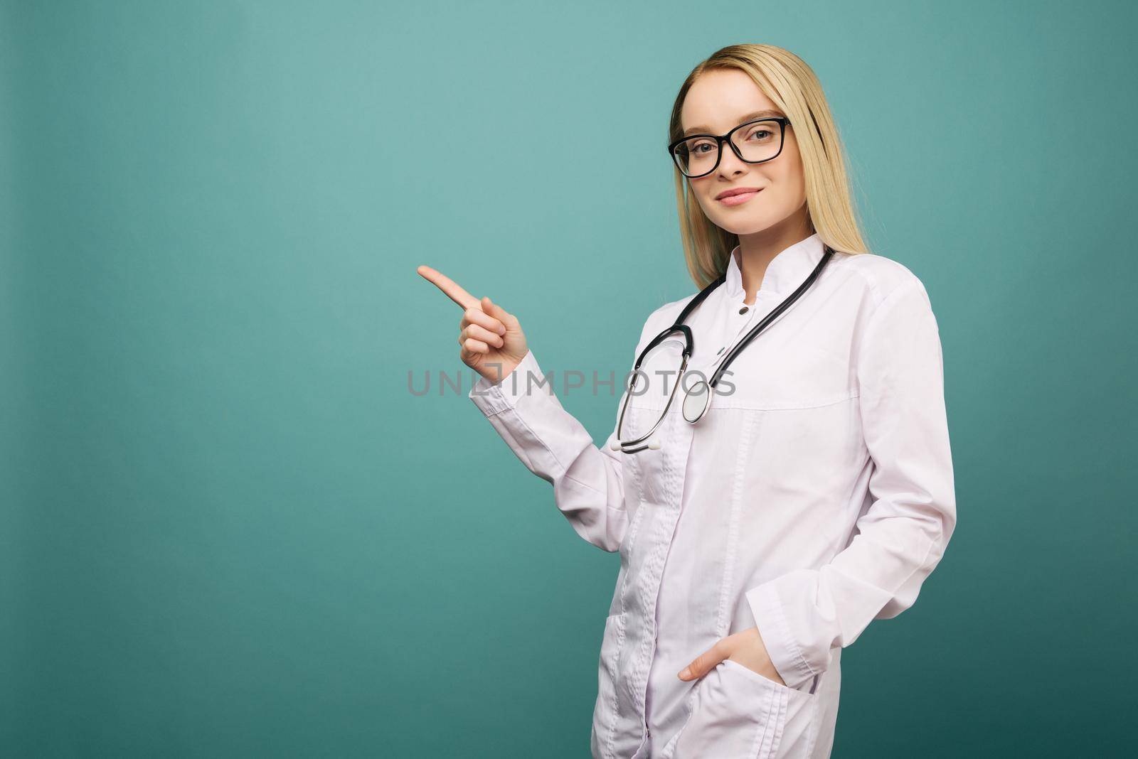 Smiling young medical doctor woman with stethoscope pointing on copyspase. Isolated over blue background.