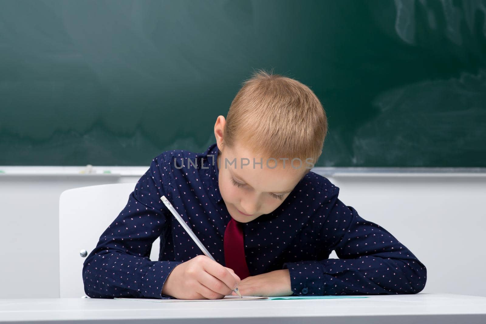 Schoolboy writing in notebook. School student in blue shirt and tie sitting at desk on background of blackboard in classroom. Back to school, education concept