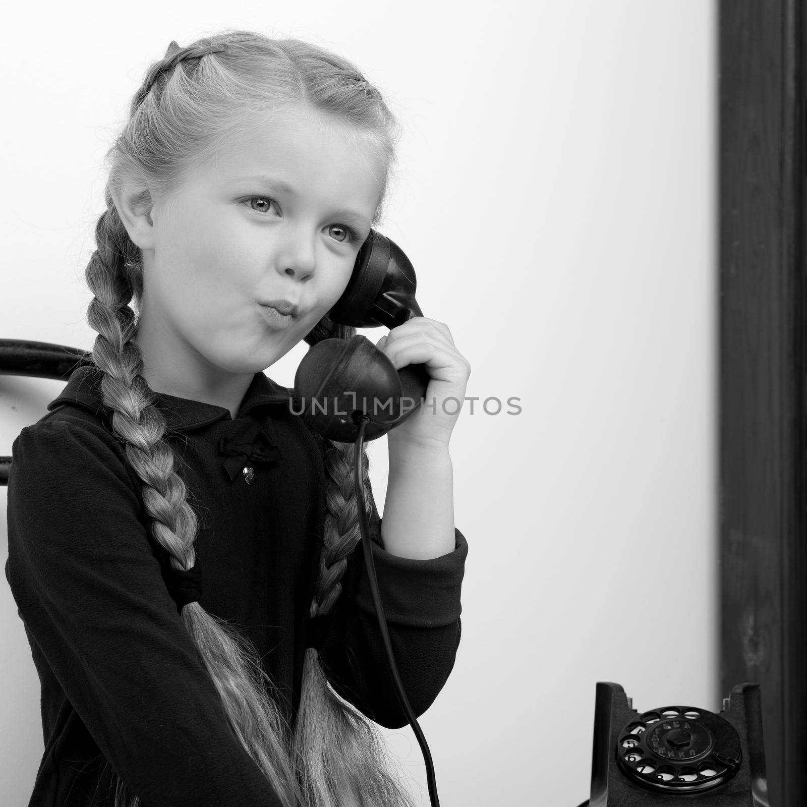 Smiling girl talking by old phone. Black and white shot of lovely kid sitting on chair in vintage room interior. Cute six years old kid speaking on vintage telephone