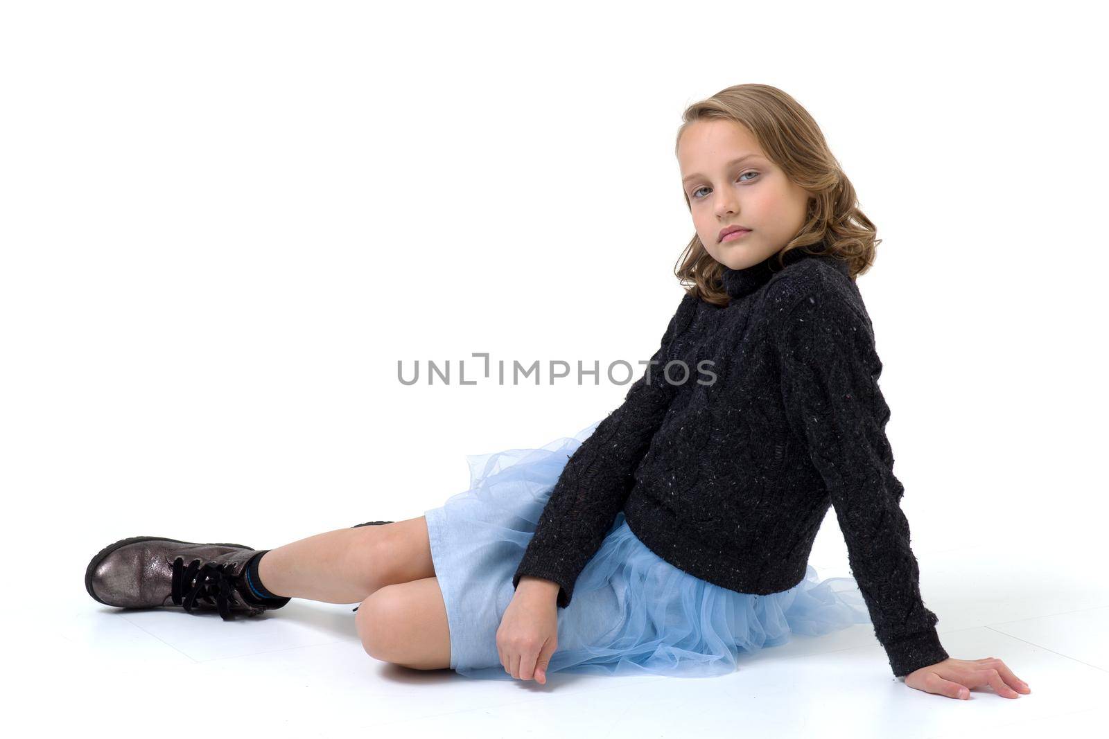 A beautiful girl in a black jumper and a blue fluffy skirt sits on the floor. Pretty blonde woman in fashionable clothes posing in studio on white background