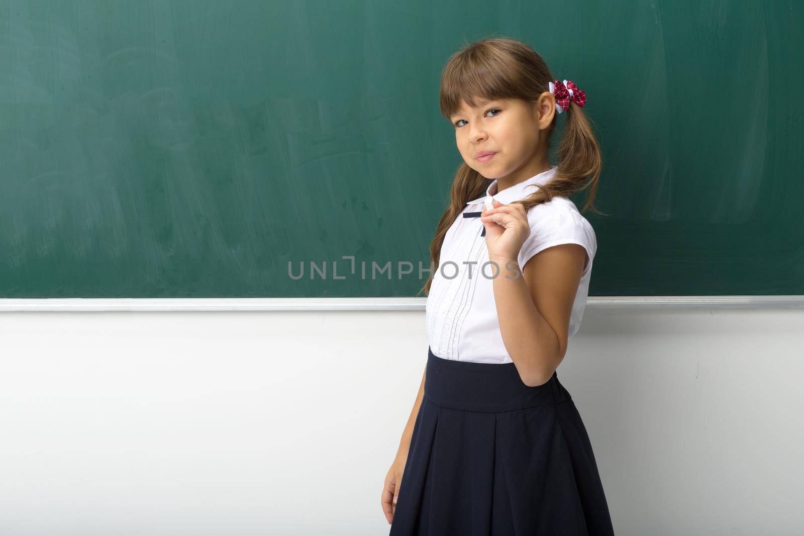 Pretty girl posing at blackboard. Portrait of beautiful schoolgirl in school uniform standing at green chalkboard with piece of chalk in her hand and looking at camera. School and education concept