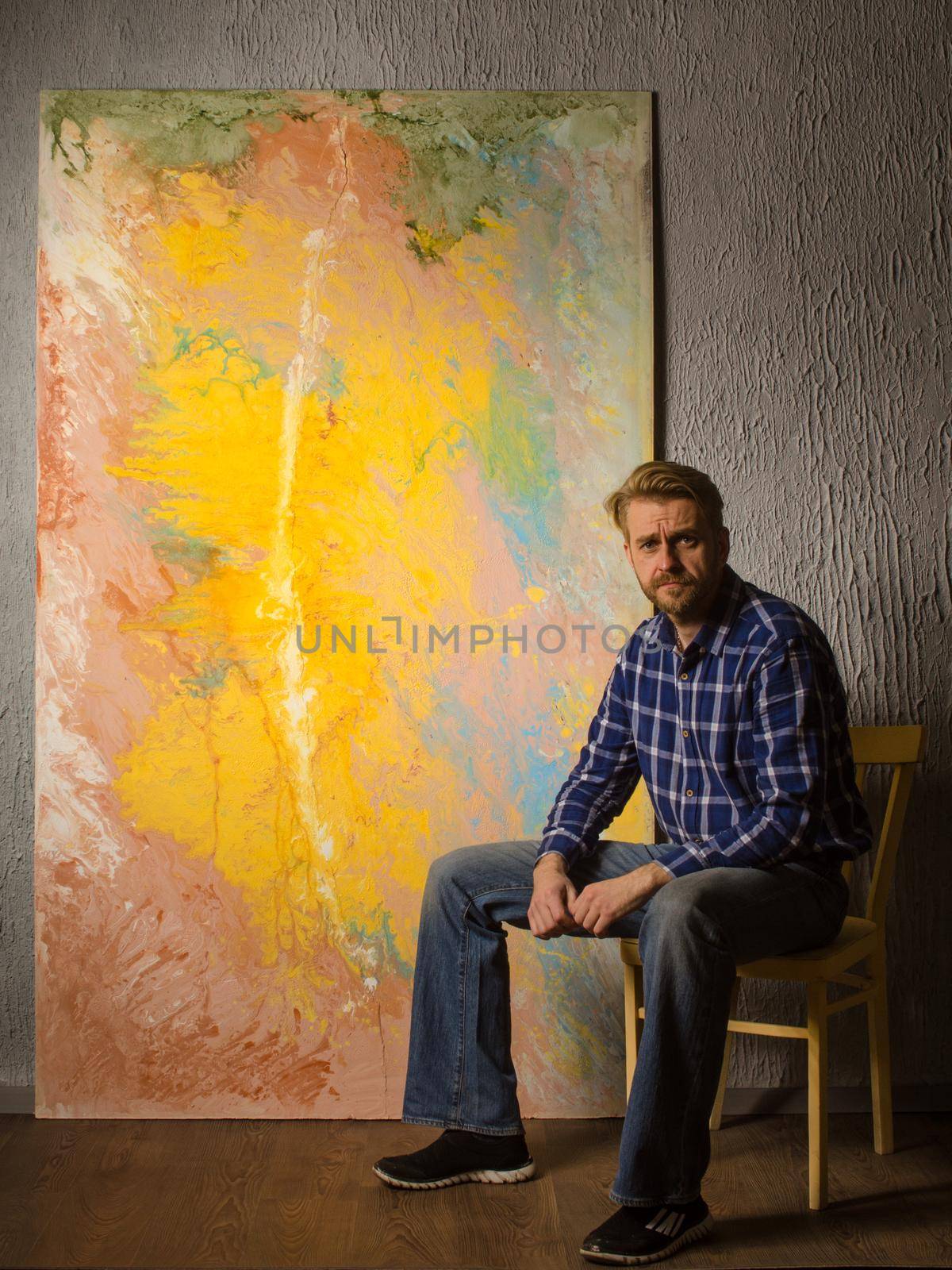 Portrait Of Male Artist sits next to his abstract painting in the Studio.