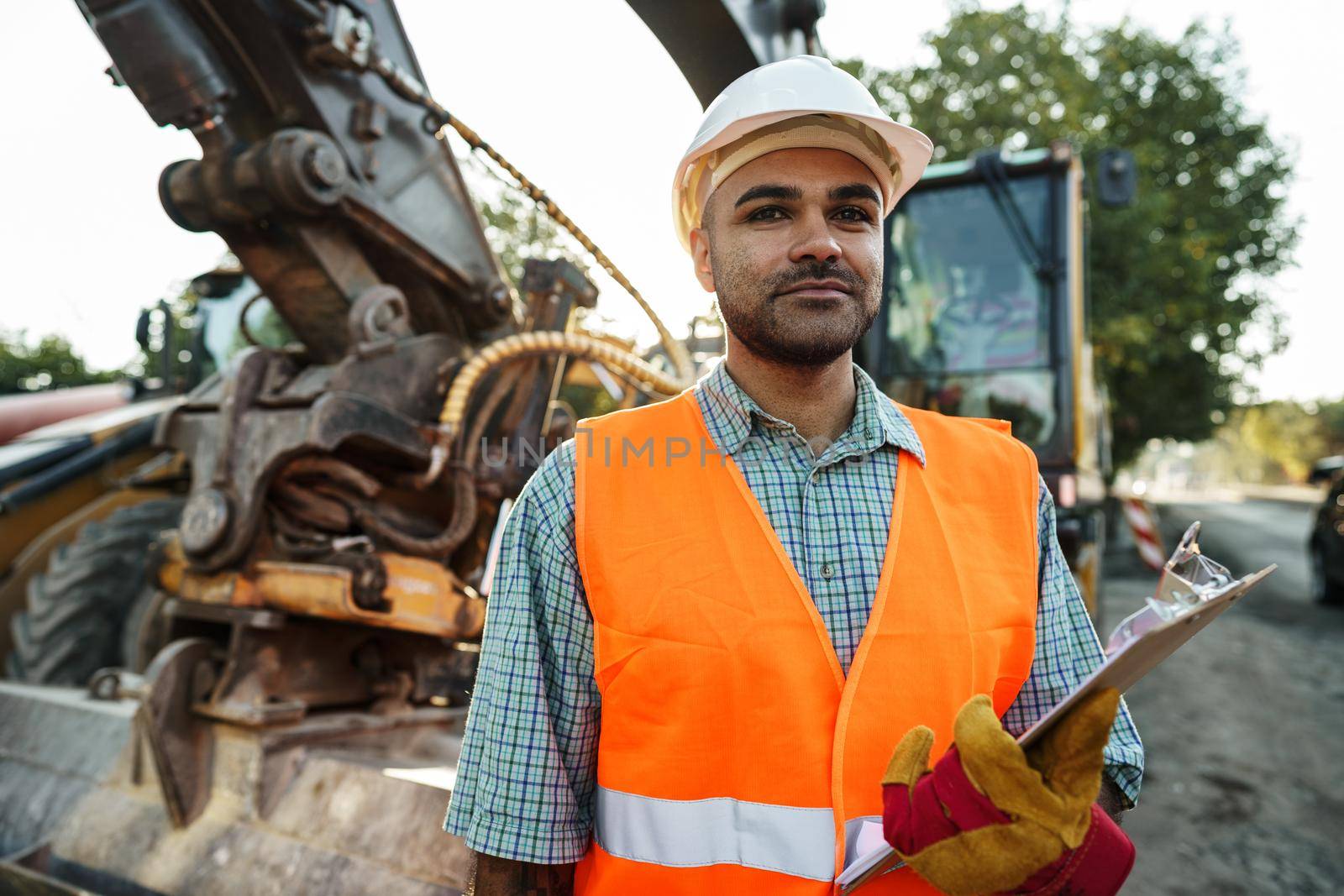 Young man engineer in workwear standing in construction site with clipboard, close up portrait